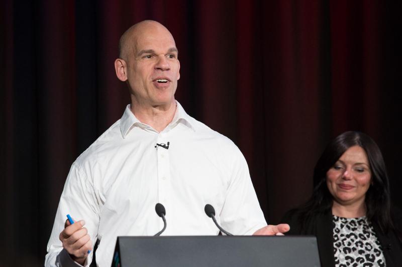 Paul Shetler presenting at a lectern with Jane Speechley in the background.