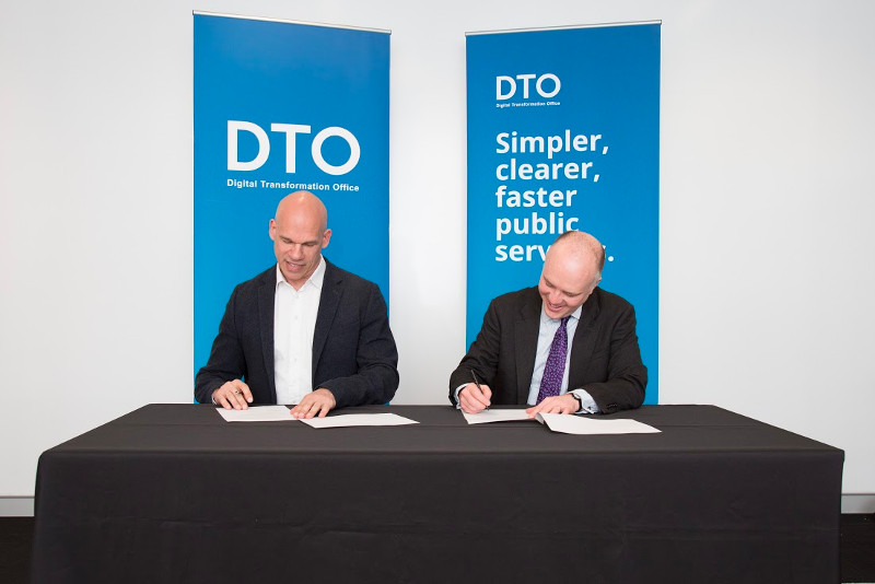 DTO CEO Paul Shetler and the Chief Technology Officer HM Government signing the MOU in front of DTO banners.